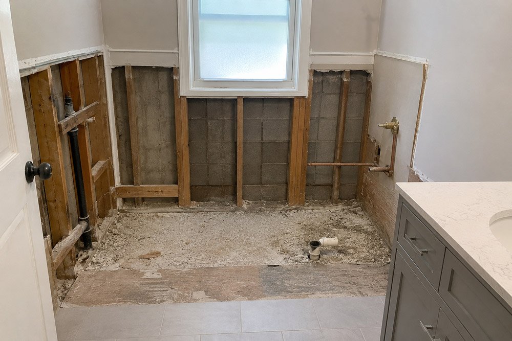 Bathroom renovation: exposed studs, concrete blocks, and plumbing with a window above; gray vanity on the right.